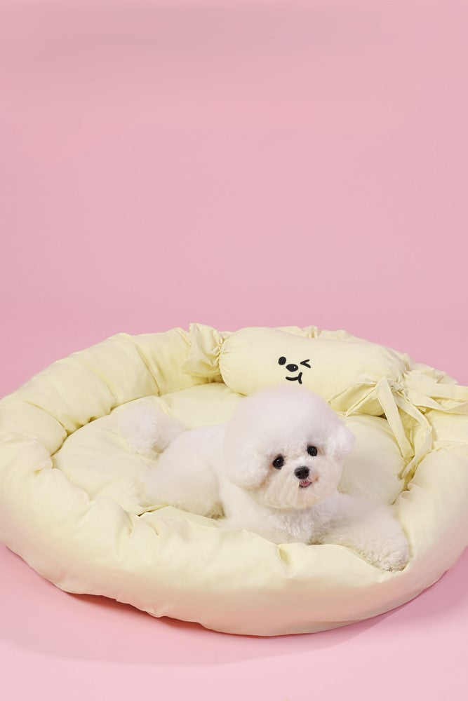 Small white dog lying on a yellow pet bed with a pink background